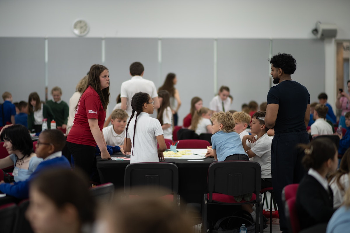 A busy workshop room with young people sitting at tables working on projects, with adult facilitators supporting the sessions during the Deliver You Hack event